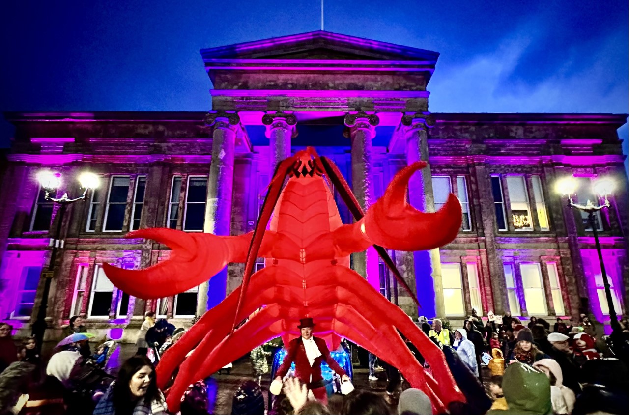The Town Hall is lit up with blue and purple lights, the sky is dark behind, and there is a large inflatable lobster which is lit up red, infront of the town Town Hall